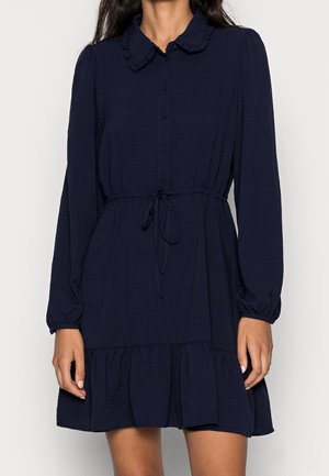 Femme portant une robe bleu marine mi-longue à manches longues avec des boutons, un col à volants, une taille nouée et un ourlet à volants.
