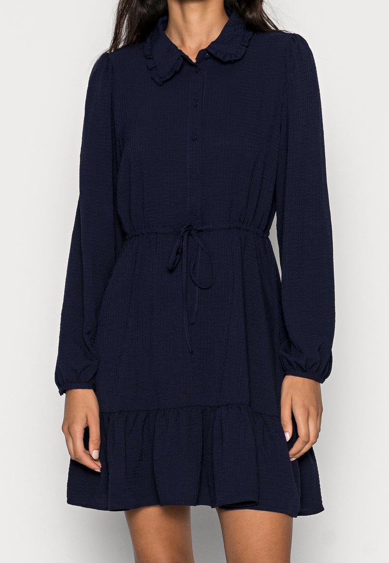 Femme portant une robe bleu marine mi-longue à manches longues avec des boutons, un col à volants, une taille nouée et un ourlet à volants.