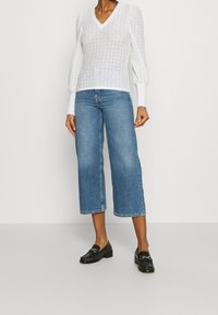 Bílý strukturovaný top s dlouhým rukávem a výstřihem do V, kombinovaný se světle modrými kalhotami s širokými nohavicemi a černými polobotkami se zdobnými detaily.