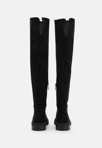 Cizme negre din suede, peste genunchi, cu o formă conică, având un design cu slit în partea de sus și fermoare pe laturile exterioare. Talpă din cauciuc plat.