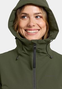 Glimlachende vrouw met blauwe ogen die een olijfgroene hoodie draagt, tot aan de kin dichtgeritst, tegen een effen achtergrond.
