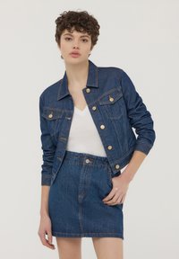 Giacca di jeans con chiusura a bottoni, due tasche sul petto e una silhouette aderente. Indossata su una maglietta bianca con scollo a V e abbinata a una gonna in denim coordinata.