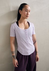 Haut lilas à côtes avec manches courtes, devant à boutons et ourlet arrondi, associé à un pantalon violet foncé. Tissu lisse et design ajusté.