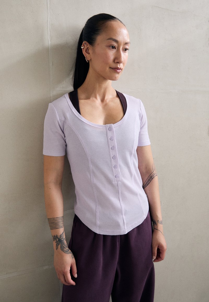 Haut lilas à côtes avec manches courtes, devant à boutons et ourlet arrondi, associé à un pantalon violet foncé. Tissu lisse et design ajusté.