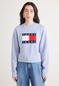 Lavendelkleurige gebreide trui met geribde mouwen en zoom. Kenmerkt zich door een centraal logo in marineblauw, wit en rood. Gecombineerd met lichtblauwe jeans.