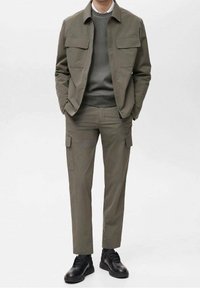 Veste cargo vert olive et pantalon assorti, tous deux en tissu lisse ; dotés de grandes poches, associés à des chaussures noires à lacets.