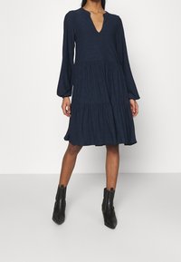 Robe bleu marine avec un décolleté en V, de longues manches bouffantes et une jupe à volants. Le tissu possède un motif texturé subtil. Assortie à des bottines noires.