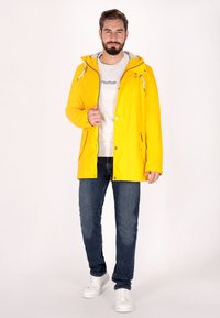 Geel waterdichte jas met een rits aan de voorkant en een capuchon, met contrasterende blauwe hardware en twee zijzakken, gedragen over een grijze sweatshirt en jeans.