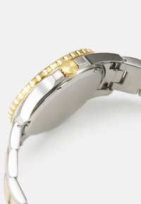 Montre en argent et doré avec un bracelet en acier lisse, un boîtier rond, une lunette dorée cannelée et une couronne dorée proéminente sur le côté.