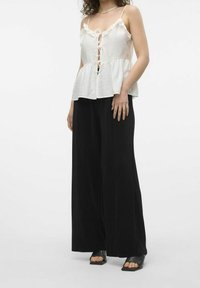 Bílý texturovaný peplum top s volánkovými okraji a středovým zavazováním, v kombinaci s širokými černými kalhotami a černými plochými sandály.