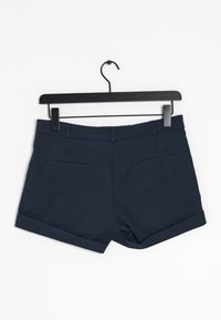 Námořnicky modré šortky z měkké tkaniny s čistým designem, dvěma zadními kapsami a zatočenými lemy. Visí na černém ramínku s klipem.