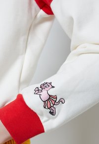 Bílá mikina s červenými žebrovanými manžetami; má na rukávu vyšívanou růžovou kočku v sukni, která vykazuje jemné detaily a živé barvy.