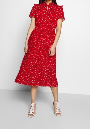 Femme portant une robe midi rouge à pois abstraits blancs, manches courtes et col à nœud, associée à des sandales blanches à talons.