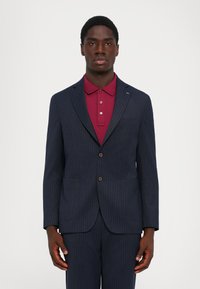 Uomo in un abito a righe blu navy con una maglietta polo bordeaux, in piedi davanti a uno sfondo bianco semplice, rivolto in avanti.