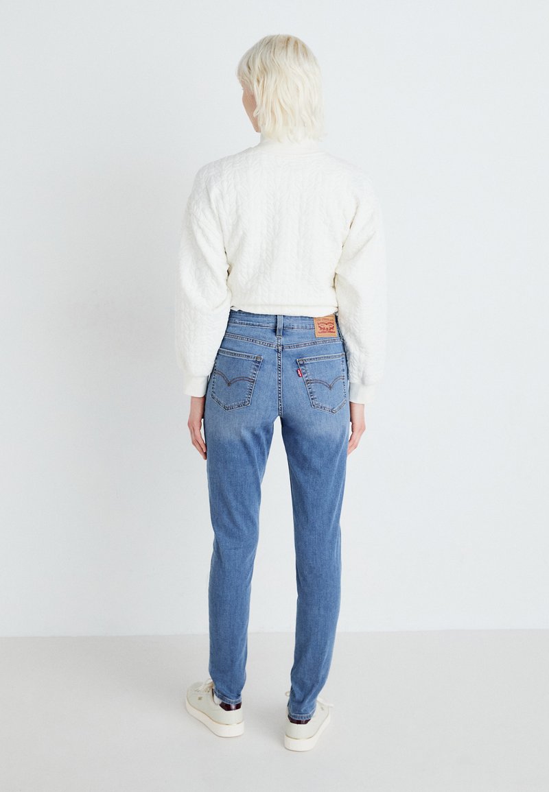 Hellblaue Jeans mit hoher Taille, ausgestattet mit zwei Gesäßtaschen und einem Leder-Brandpatch. Das Modell trägt einen strukturierten weißen Pullover.