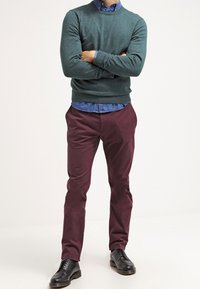 Maglione verde a girocollo, camicia blu con colletto, pantaloni bordeaux e scarpe in pelle nere. Il maglione presenta polsini e orlo a coste.