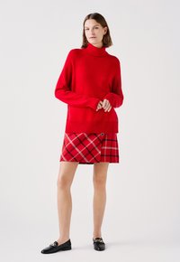 Czerwony sweter z golfem o żebrowanym wykończeniu, w zestawieniu z czerwoną spódnicą w kratę z białymi i czarnymi akcentami, uzupełniony czarnymi mokasynami.