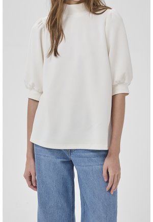 Kobieta ubrana w białą bluzkę z bufkami i wysokim kołnierzem oraz jasnoniebieskie dżinsy, stojąca na jednolitym tle.