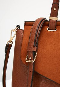 Sac à main en cuir et daim marron avec boucles dorées et bandoulière amovible sur fond blanc.