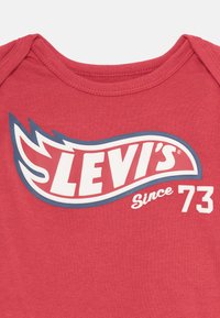 Material textil roșu cu logo-ul Levi's în alb și albastru, având un design stilizat de flacără și textul "Din 73" în zona superioară a pieptului.