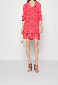 Vestido de encaje coral con dobladillo en forma de concha, escote en V y mangas tres cuartos. Combinado con sandalias de tacón altas en marrón y verde y un bolso pequeño.