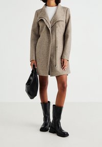 Beige villasekoitepäällinen takki, jossa on suuri kaulus ja vetoketju, täydentää musta nahkalaukku ja polven yli ulottuvat mustat korkosaappaat.