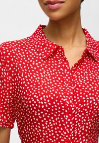 Chemise rouge à motif blanc moucheté, manches courtes et col pointu. Fermeture à boutons sur le devant.