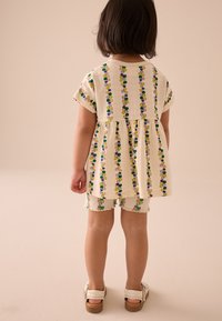 Vestito in tessuto con vita arricciata, colore crema, e motivi floreali verticali in viola, giallo e verde, abbinato a pantaloncini e sandali coordinati.