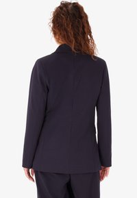 Blazer navy con vestibilità su misura, dotato di uno spacco posteriore singolo, maniche lunghe e una consistenza liscia. Design minimalista senza hardware visibile.