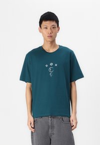 T-shirt in cotone turchese con maniche corte; presenta un design grafico celestiale bianco al centro del petto. Veste casual e colletto rotondo.