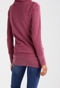 Persona che indossa un hoodie a maniche lunghe color maroon e jeans blu, vista di spalle su uno sfondo chiaro e uniforme.