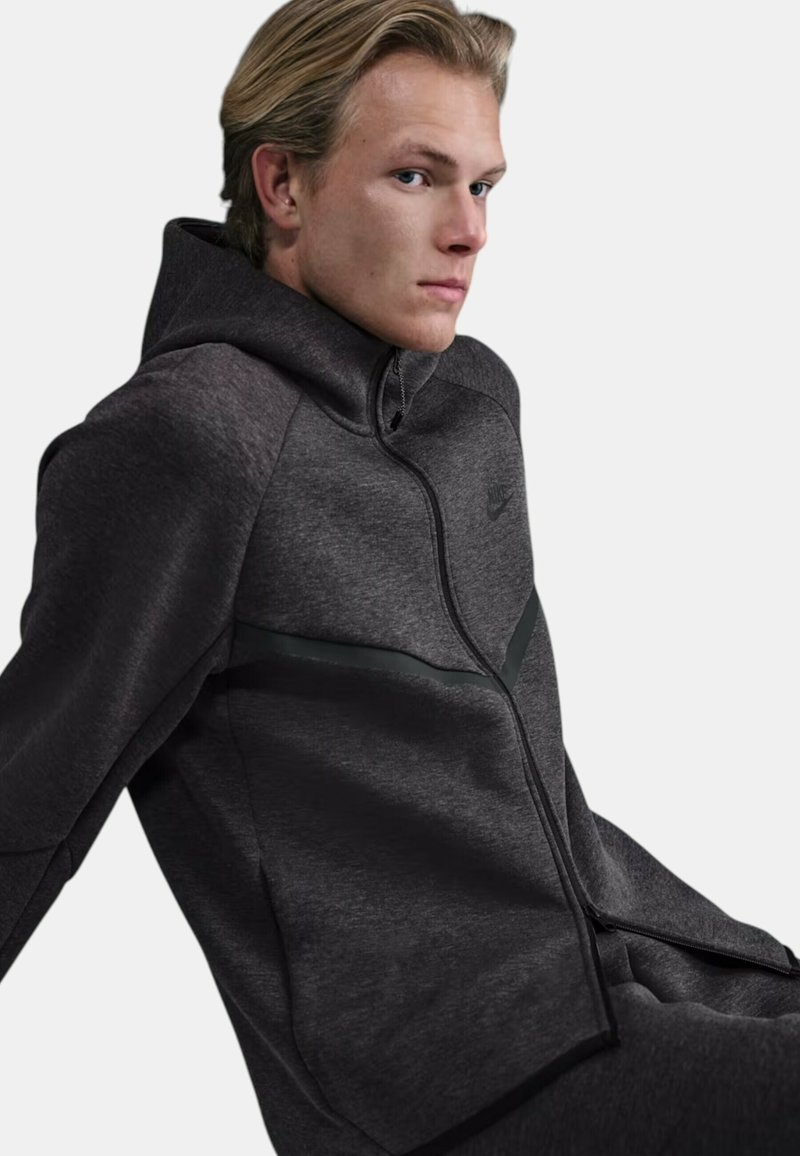 Mörkgrå zip-up-hoodie i texturerat tyg med en kontrasterande svart rand över bröstet och en figursydd huva. Nike-logotyp finns.