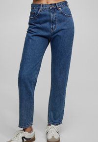 Jeans z wysokim stanem w niebieskim kolorze, z prostymi nogawkami, z pięcioma kieszeniami, przeszyciami oraz klasycznym zapięciem na guziki. Dobrane z białymi trampkami.