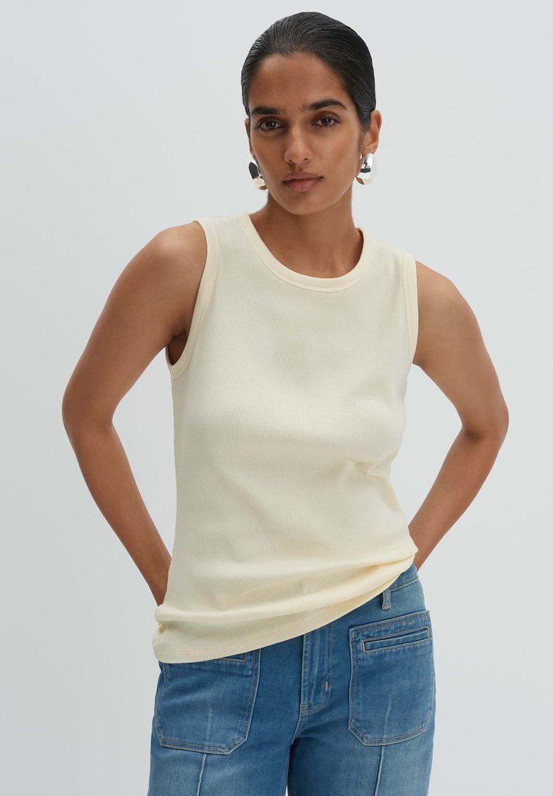 Donna con capelli scuri, indossa una canottiera color crema senza maniche e jeans blu, in piedi con le mani dietro la schiena davanti a uno sfondo chiaro e uniforme.