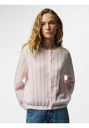 Jeune femme aux longs cheveux blonds portant un cardigan en maille rose clair boutonné et un jean bleu sur un fond blanc uni.