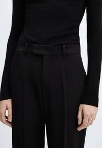 Pantalons noirs ajustés avec une texture lisse, dotés d'une ceinture, de plis et d'une coupe droite. La taille présente des détails de couture subtils.