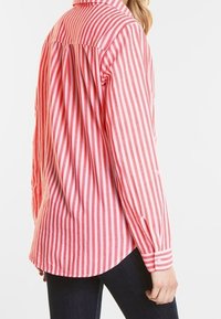 Chemise à rayures rouges et blanches à boutons, avec col, manches longues et ourlet arrondi. Tissu texturé avec détails de couture visibles.