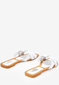 Sandalias planas blancas con tiras cruzadas, hechas de material sintético. Presentan una suela rectangular de madera y un logo impreso en la plantilla.