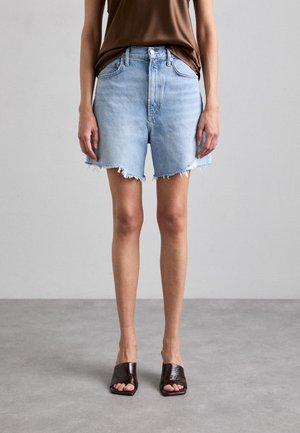 Persona que lleva pantalones cortos vaqueros desgastados de color azul claro, top marrón y sandalias mule de punta abierta marrón oscuro, de pie sobre un suelo gris.