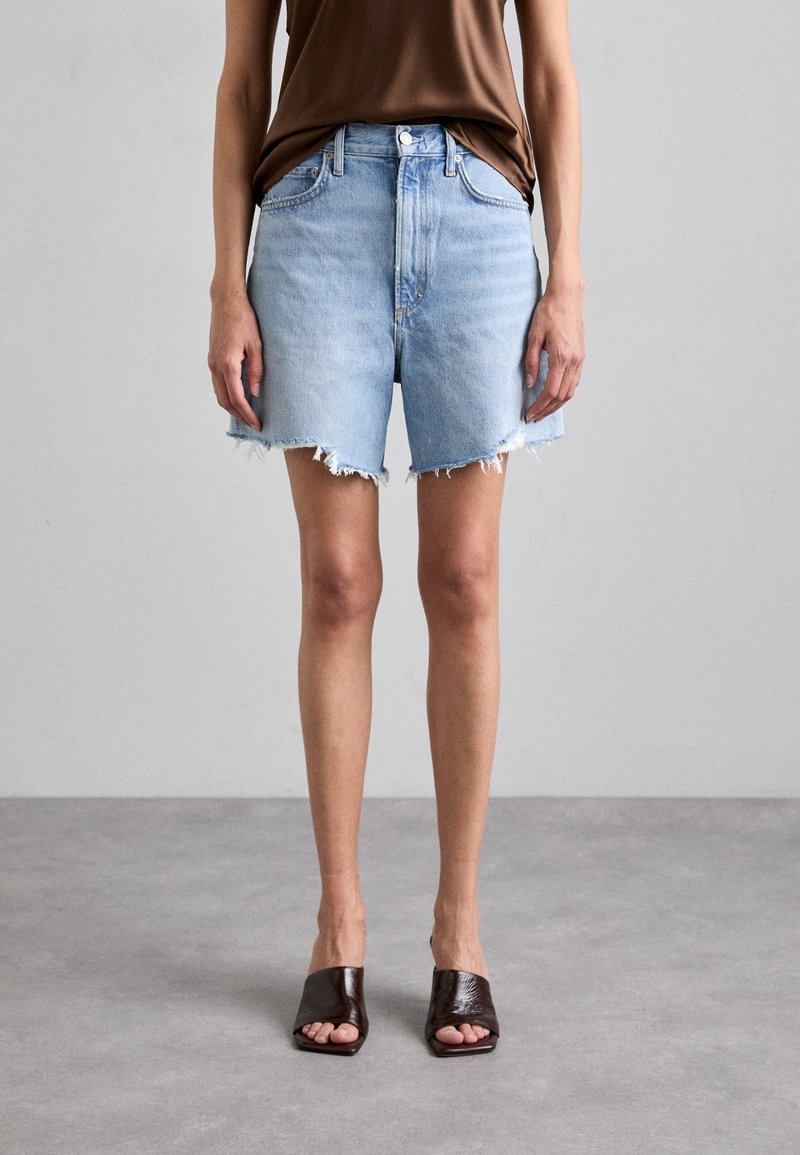 Personne portant un short en jean bleu clair effiloché, un haut marron et des sandales mule marron foncé à bout ouvert, debout sur un sol gris.