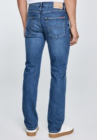 Hátsó nézet egy olyan személyről, aki egyenes szárú kék farmert és fehér sportcipőt visel, egy egyszerű világos háttér előtt állva.