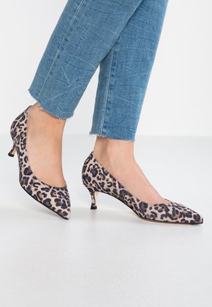 Pieds portant des escarpins à talons kitten imprimé léopard, associés à un jean bleu raccourci avec des ourlets effilochés, debout sur un sol blanc.