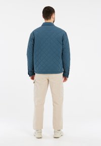 Gequilted blauwe jas met een kraag, voorzien van een gestructureerd ruitpatroon. Gepaard met beige cargobroeken en lichte schoenen.