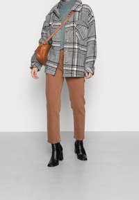 Veste-chemise en laine à carreaux gris avec de gros boutons, portée sur un pull bleu côtelé et un pantalon marron, associée à des bottines noires à talons et un sac marron.