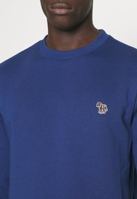 Sudadera azul con cuello redondo y un pequeño logo de animal bordado multicolor en el lado izquierdo del pecho. Textura suave, corte informal.