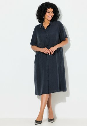 Marineblauwe blouse jurk met korte mouwen, lichtgewicht textuur stof, knielang en zijzakken. Gecombineerd met zwarte puntige schoenen.