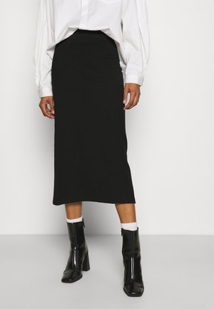 Schwarzer, gerippter Midirock aus weichem Material, mit figurbetontem Schnitt und glatter Textur. Kombiniert mit glänzenden schwarzen Stiefeletten.