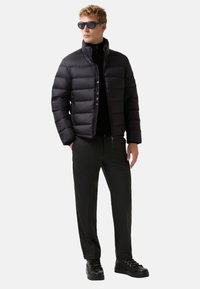 Mand iført sort pufferjakke, sort turtleneck, mørkegrå bukser, sorte sneakers og runde sorte solbriller, stående med hænderne i lommerne.