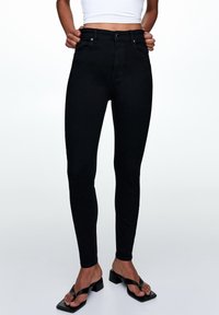 Jean noir taille haute à coupe slim, avec fermeture par boutons et poches avant, assorti à des sandales noires à brides.