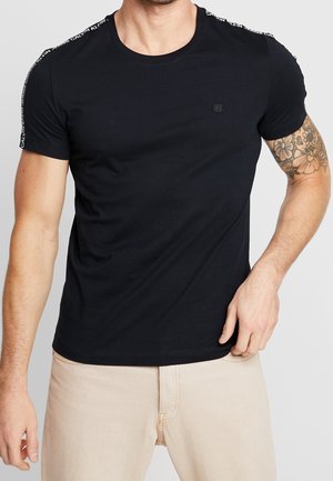 T-shirt noir en coton à manches courtes, avec une marque blanche sur les épaules et un petit logo sur la poitrine. Design doux et ajusté.